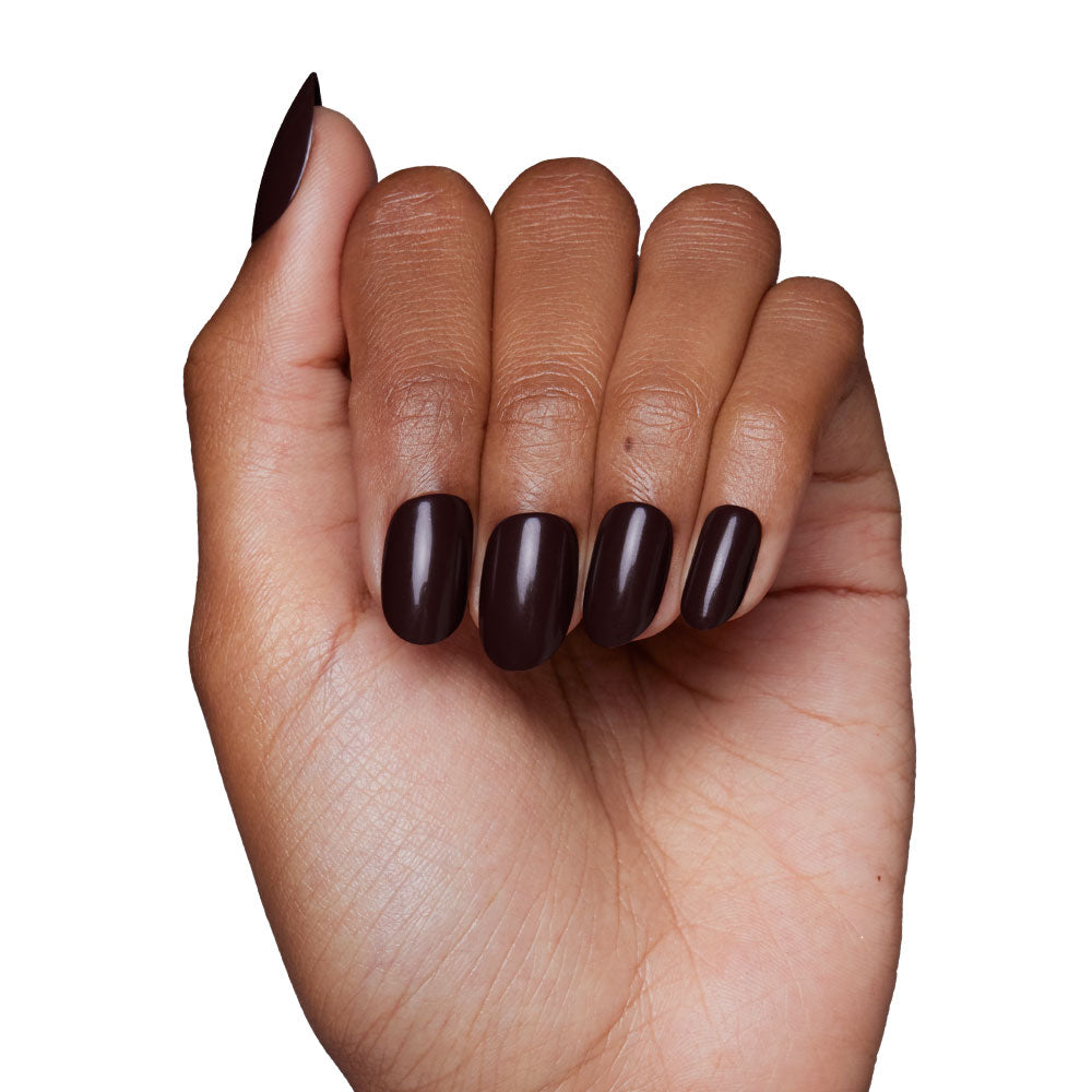 A hand with medium-brown skin displays the Bark Brown nail set by glamnetic, neatly manicured and painted in dark brown polish, shown against a white background.