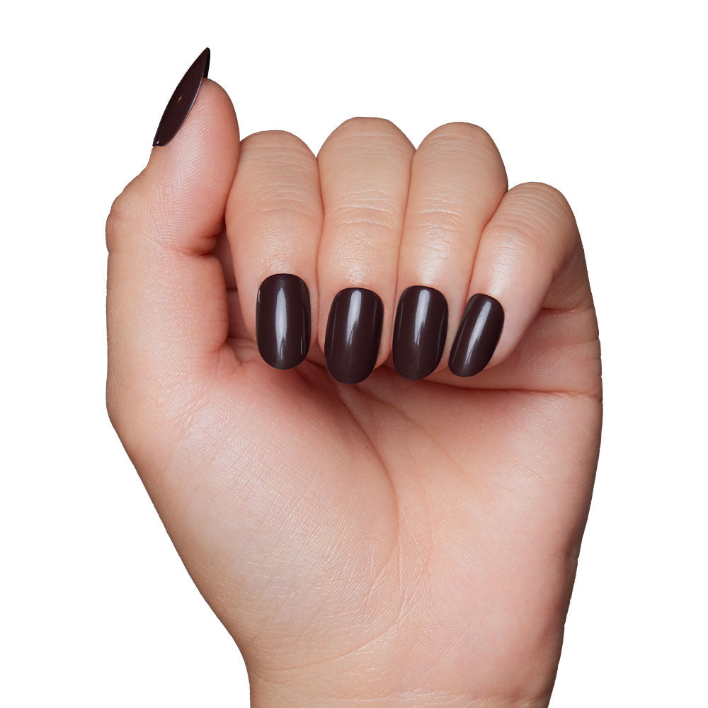A hand with medium-length oval nails painted in glamnetics Bark Brown, a glossy dark brown polish, captures cozy vibes of a brown nail set against a white background.