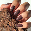 A hand with short, neatly manicured nails painted in glamnetics Bark Brown glossy set holds a piece of textured bark.