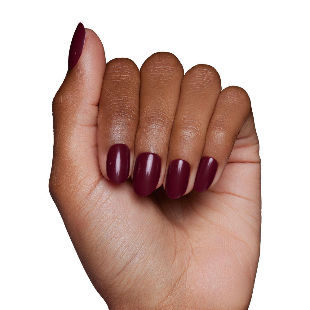 A hand with medium brown skin tone displays oval-shaped glamnetic Berry Maroon press-on nails, the glossy maroon color standing out as fingers curl inward against a crisp white background.