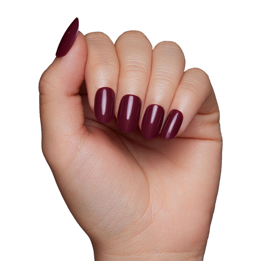 A hand with glamnetics Berry Maroon press-on nails, featuring medium-length oval tips in a glossy dark burgundy shade, is displayed against a plain white background.