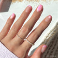 A hand with manicured nails features glamnetic’s Blush Dunes press-on set, showcasing seashell designs, soft pink chrome tips, and a starfish accent. The look is complemented by pearl and silver rings against a softly lit background.