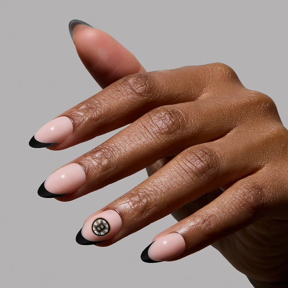 The glamnetic Boston Bruins Press-On Nail Set features medium almond-shaped nails with a nude base, black French tips, and a gold and black circular accent on the ring finger for a subtle fandom look.