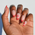 A hand with almond-shaped nails features glamnetics Boston Red Sox Press-On Nail Set, displaying red French tips and a small red sock graphic on the middle and ring fingers against a white background.