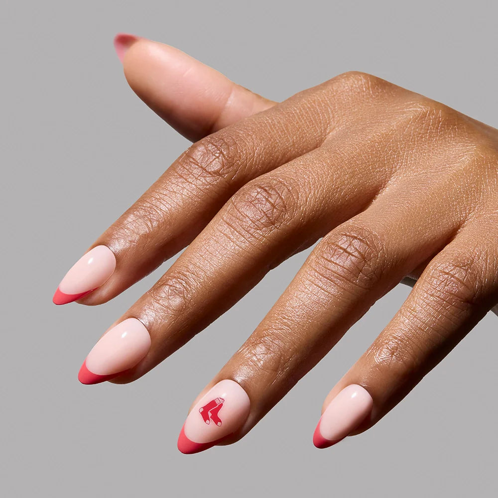 A hand with almond-shaped nails featuring a light pink base, red tips, and a small red graphic on the ring finger showcases the glamnetic Boston Red Sox Press-On Nail Set against a plain gray background.