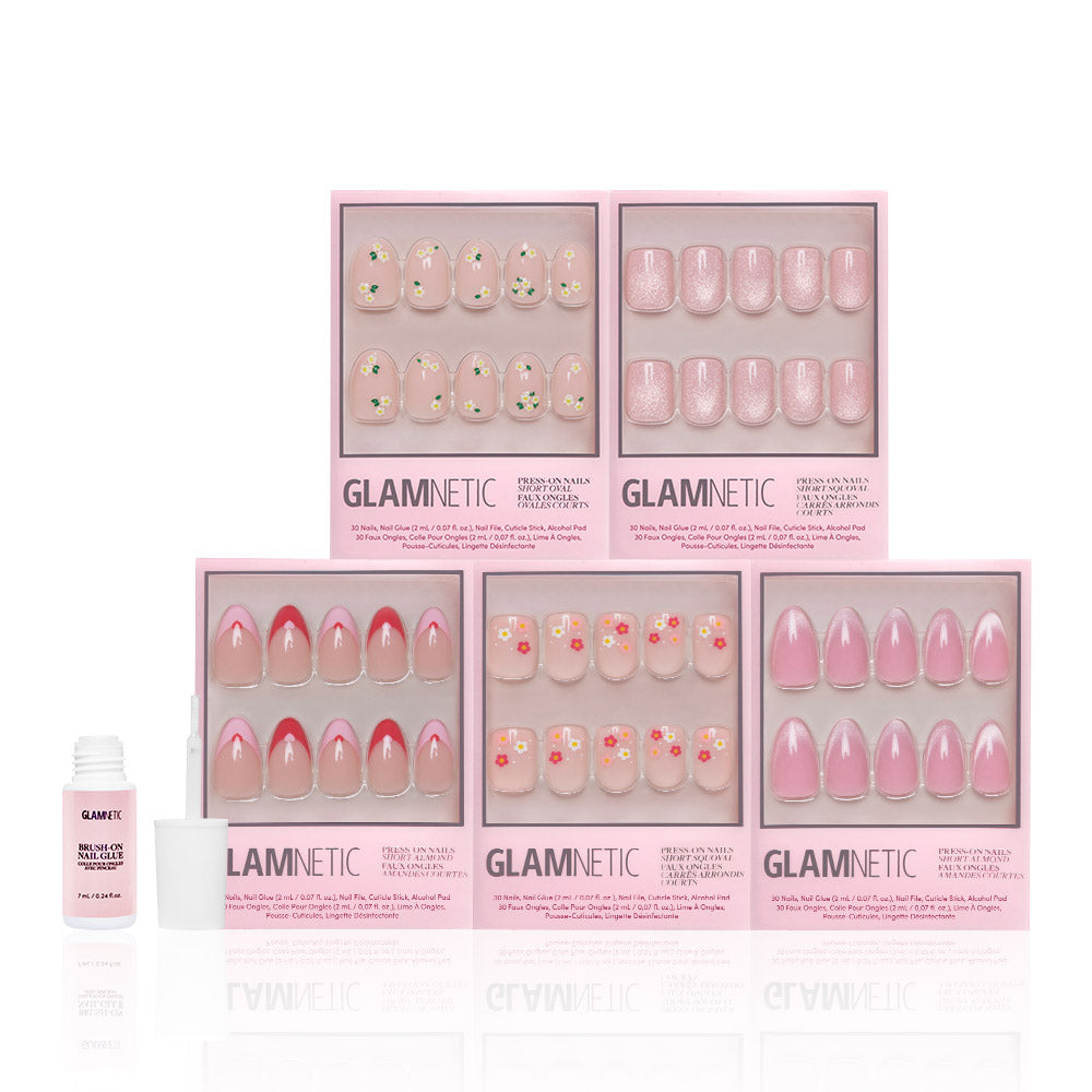 Five boxes of glamnetic Budding Romance pink-themed Glossy Nail Set press-ons, featuring short almond nails, are arranged in two rows. A small open bottle of nail glue is placed beside the boxes on a white background.