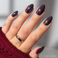 A hand with short almond-shaped nails painted in shiny, dark purple Burgundy Chrome polish by glamnetic. A silver ring and a textured dark red sweater sleeve complete the chic look of this burgundy chrome nail set.