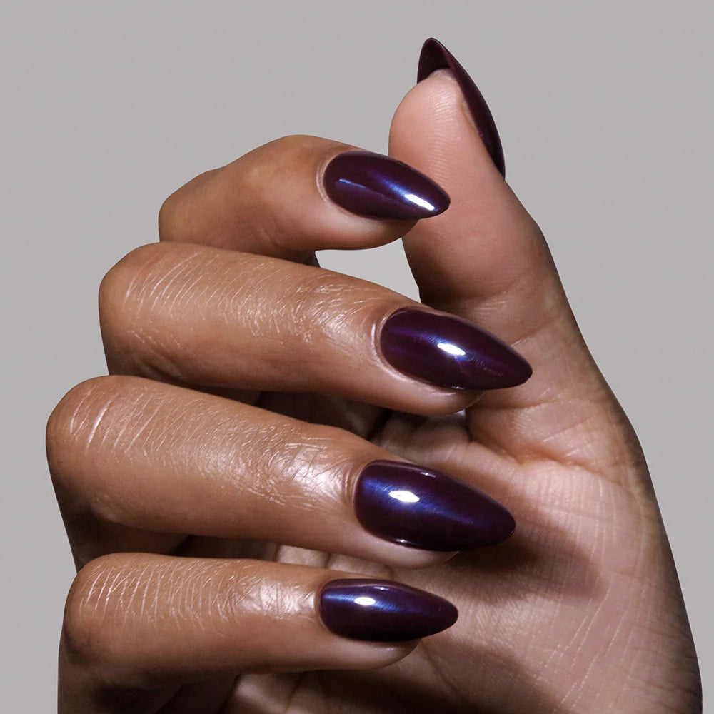 A medium brown hand displays long, pointed nails coated in glamnetic’s Burgundy Chrome polish, giving a glossy, dark purple finish for a chic and modern look.