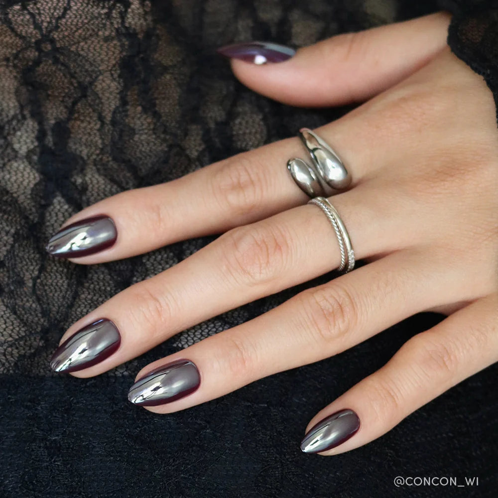 A hand with short almond glamnetic Burgundy Chrome nails features a dark, reflective polish. The person wears two silver rings, and black lace is in the background—ideal for those seeking elegant reusable press-on nails.