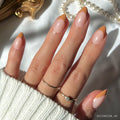 A hand with almond-shaped nails in glamnetic’s Café Crème, showcasing reusable peachy-orange French tips. Two gold rings and a white knit sweater complete the cozy look, with pearls softly blurred in the sunlit background.