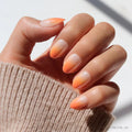 A hand with almond-shaped Candy Corn nails by glamnetic, featuring an orange ombré fade, rests on a beige ribbed sweater—a chic Halloween-ready manicure.