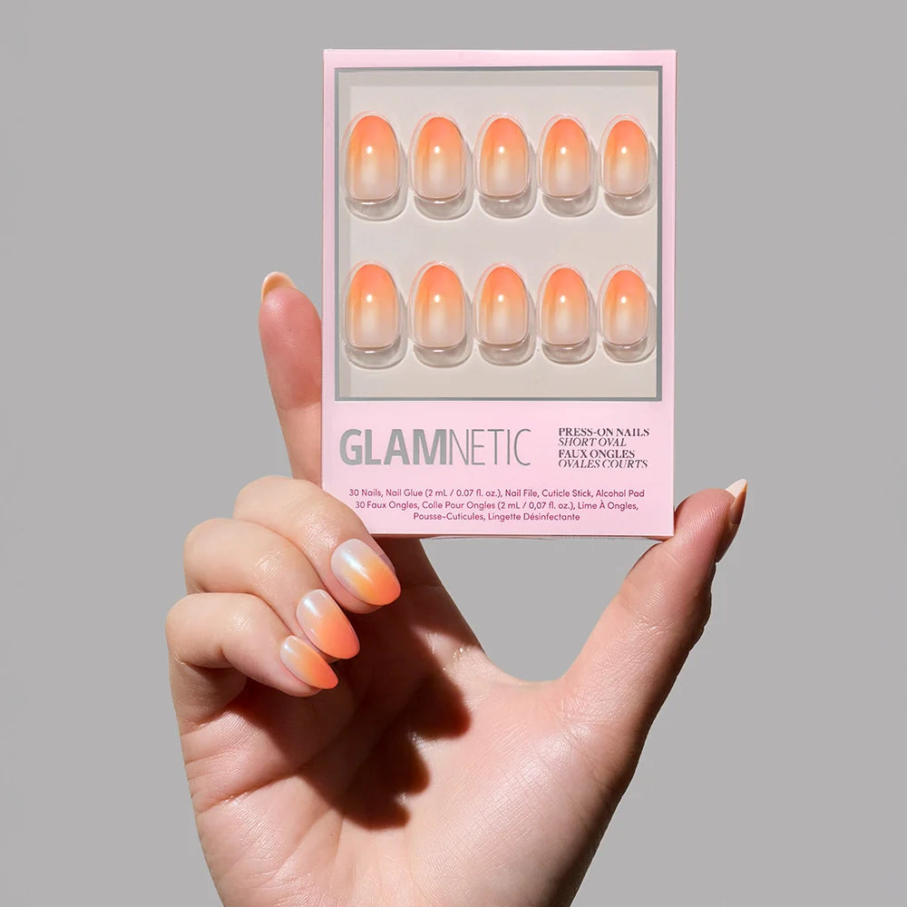 A hand holds a glamnetic Candy Corn nail set, featuring Halloween-inspired orange ombré nails that fade from peachy orange bases to pale pink tips. The light pink packaging clearly displays the product details.