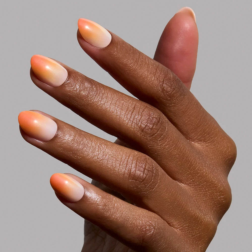 A hand with medium brown skin displays glamnetic’s Candy Corn Nail Set, featuring smooth orange and white ombré, almond-shaped nails that evoke festive Halloween style, against a plain light gray background.