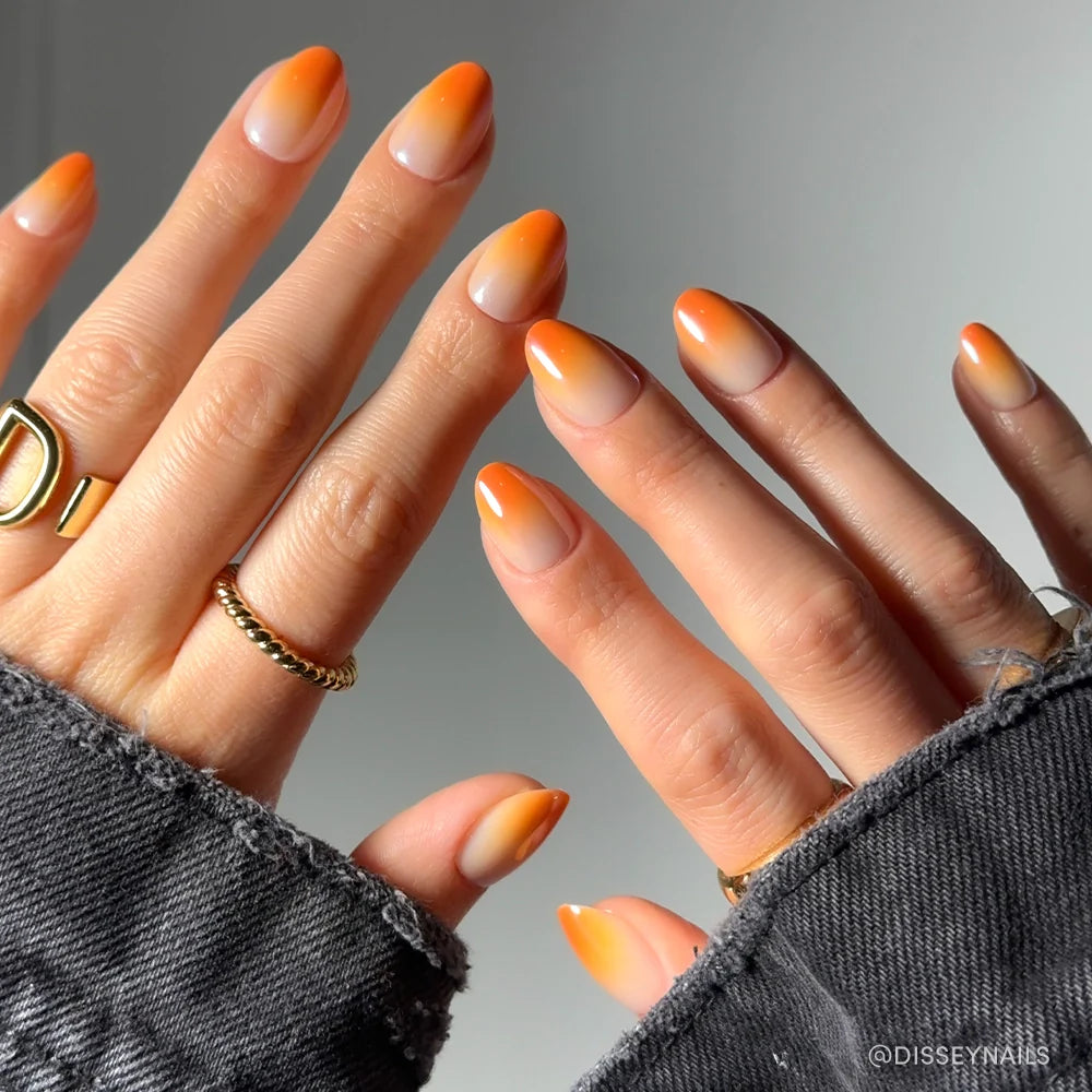 Close-up of hands with almond-shaped glamnetic Candy Corn nails, showcasing bright orange French tips and a subtle ombré effect. Gold rings and a frayed dark denim jacket complete the festive Halloween look.