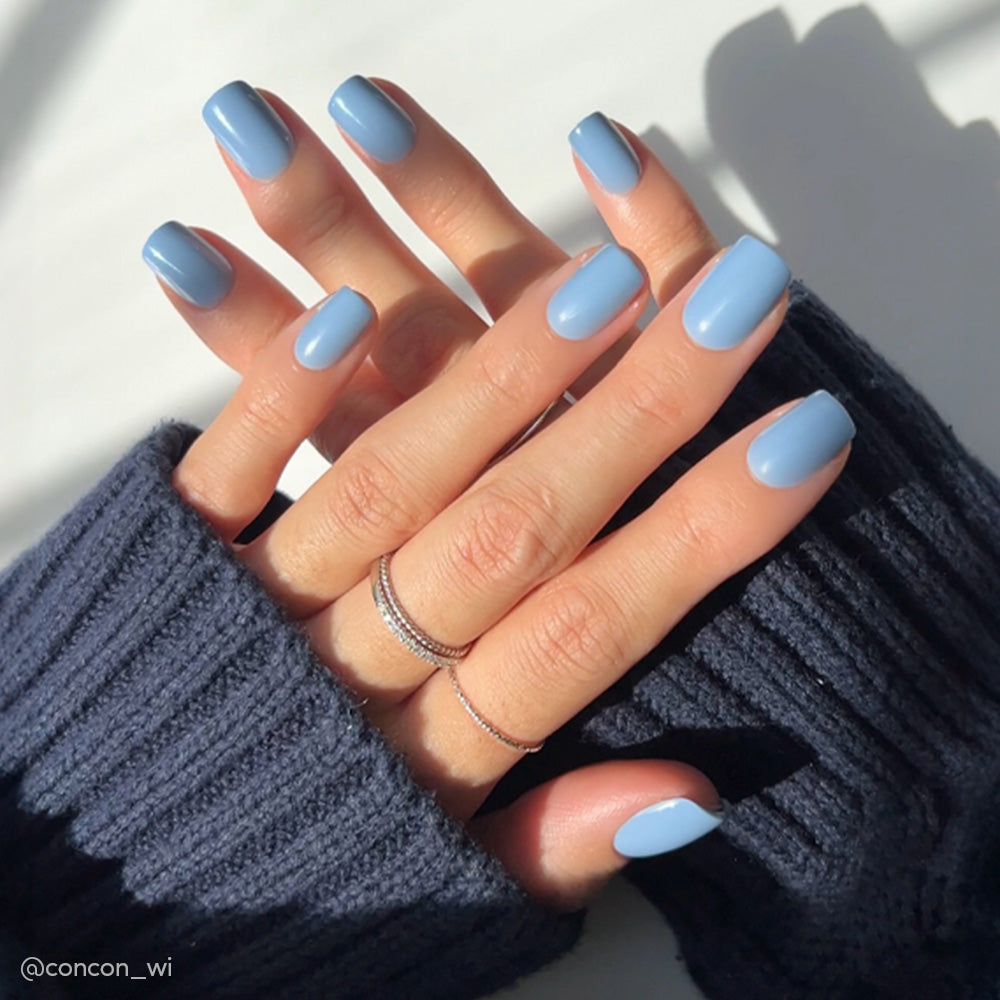 Two hands with neatly manicured short squoval nails painted in glamnetic’s Cashmere Blue rest on each other. The person wears a navy blue sweater and thin silver rings, while sunlight casts soft shadows on a light background.