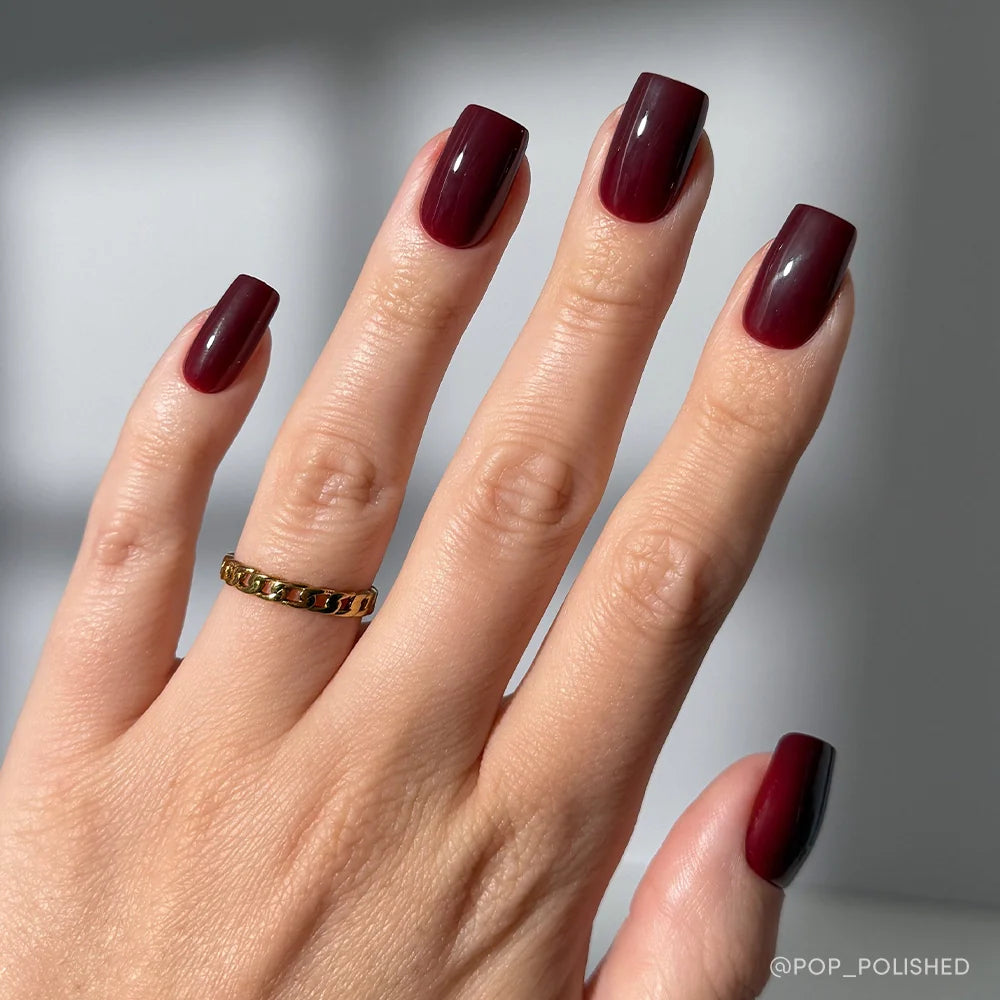 A hand with medium square Cherry Mocha nails by glamnetic in a cherry brown gloss is shown with a gold chain ring on the middle finger, set against a softly blurred background. Ideal for fans of vegan, cruelty-free style.