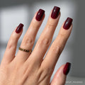 A hand with medium square Cherry Mocha nails by glamnetic in a cherry brown gloss is shown with a gold chain ring on the middle finger, set against a softly blurred background. Ideal for fans of vegan, cruelty-free style.