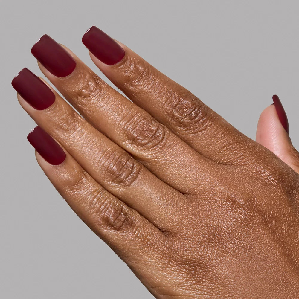 A hand with medium-dark skin tone showcases neatly manicured, medium square nails painted in glamnetics Cherry Mocha, a deep matte burgundy shade, against a plain gray background.