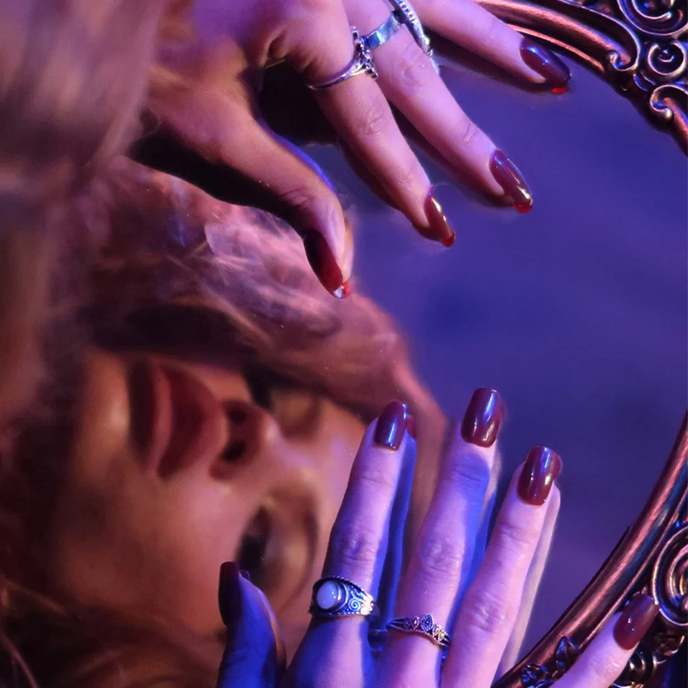 A womans face is reflected in an ornate mirror as her hands, featuring glamnetics Cherry Mocha gloss on medium square nails and adorned with rings, rest on the edge. Purple-blue lighting adds a dramatic, artistic touch.