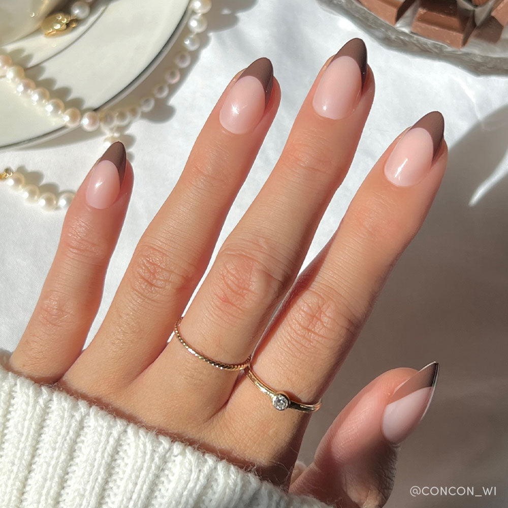A hand with short almond nails painted in glamnetics Chocolat—a pink base with dark brown French tips. Two delicate rings, a white sweater, pearls, and a dessert add elegance to the earthy-toned background.