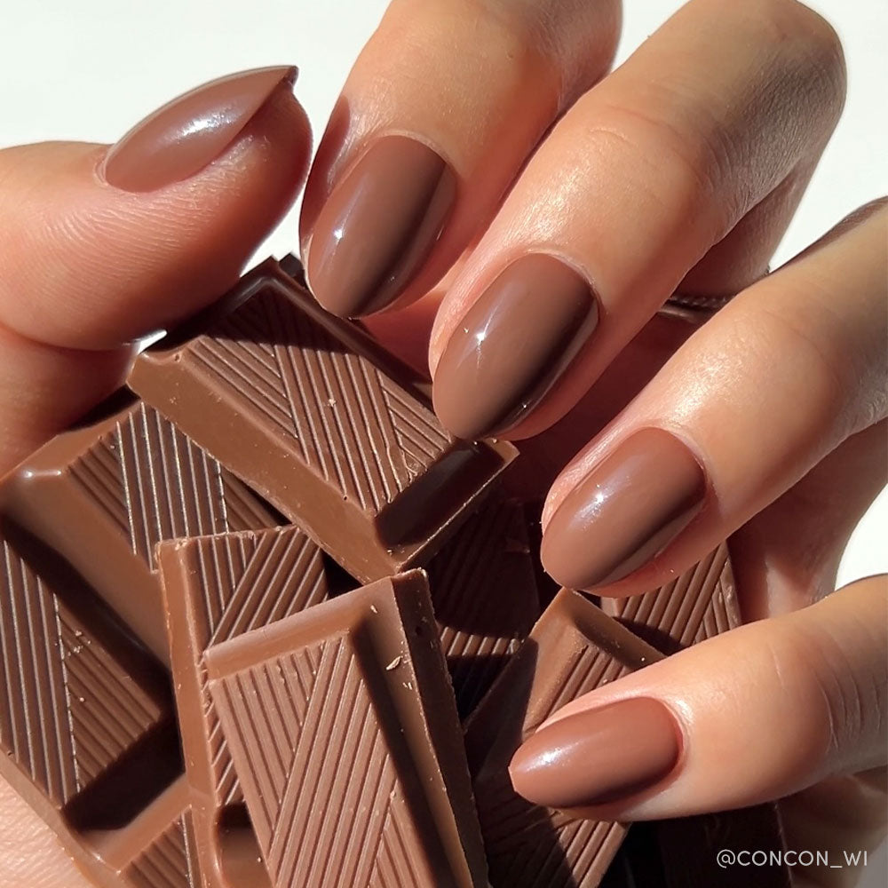 A hand with almond-shaped glamnetic Chocolate Brown Press-On Nails holds several pieces of ridged milk chocolate.