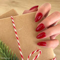 A hand with glossy Classic Red almond nails by glamnetic holds a brown paper-wrapped gift tied with red and white string; a small green pine branch peeks from the corner.