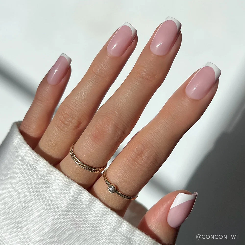 A hand with long, almond-shaped nails flaunts glamnetics Classy French mani: a modern twist on French tips featuring a light pink base and white geometric accents. Gold rings and a white cuffed sleeve complete the look.