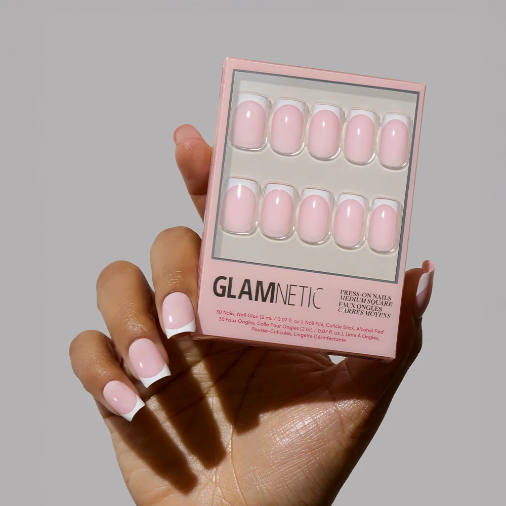 A hand with light pink, square-shaped nails holds a box of glamnetic’s Classy French press-on nails in a matching shade, showing off a trendy mani with a modern twist against a neutral background.