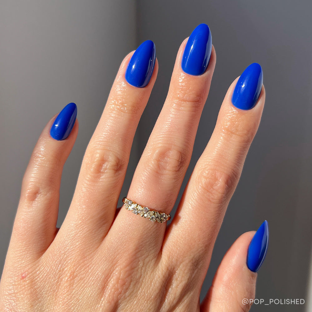 A hand with short, almond-shaped nails painted in glamnetics Cobalt Blue, perfect for summer adventures. The person wears a gold gemstone ring on the ring finger, set against a neutral, well-lit background.