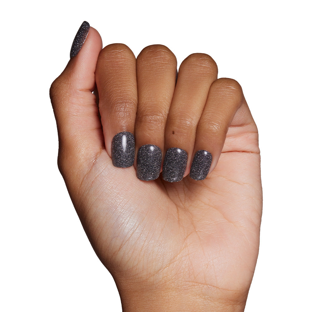 A hand with medium brown skin tone shows short squoval nails, painted in a dark gray, semi-sheer black shade inspired by the Cosmic Nail Set from glamnetic, against a white background.