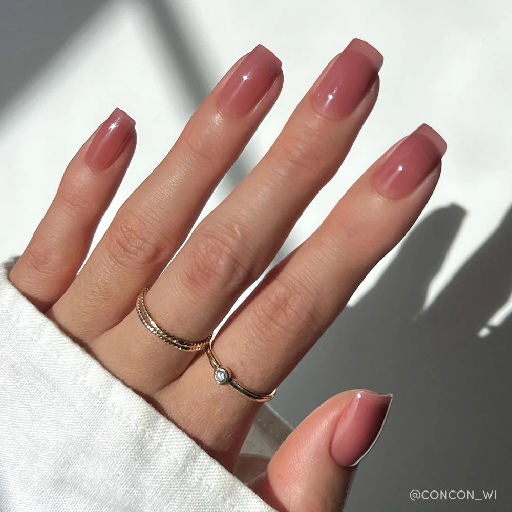 A hand with neatly manicured, square-shaped nails painted in glamnetics Dusty Pink shade, wearing two delicate gold rings—one with a small stone—set against a soft, sunlit background.