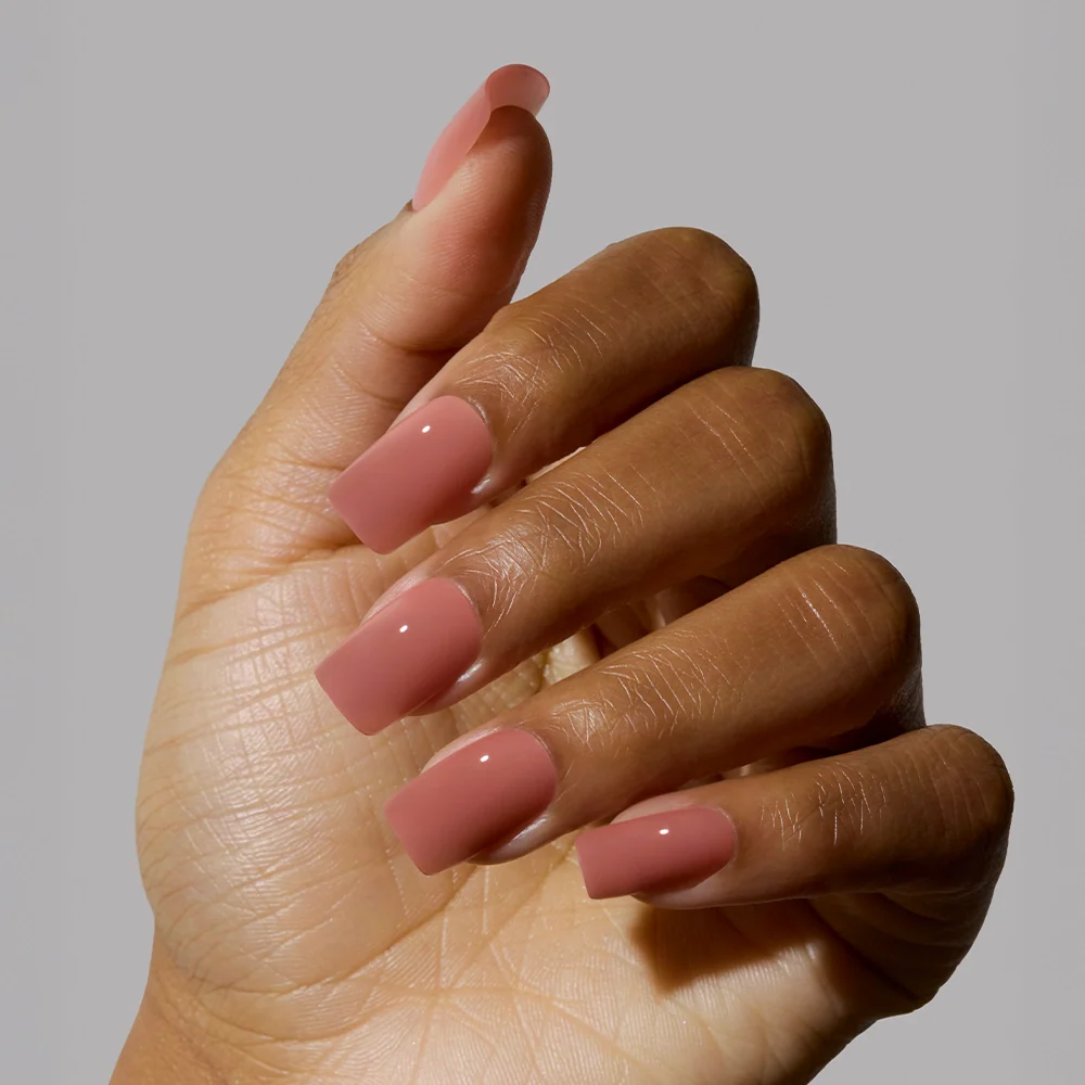 A hand with medium-dark skin tone shows long, medium square nails painted in glamnetics glossy Dusty Pink shade against a plain gray background.