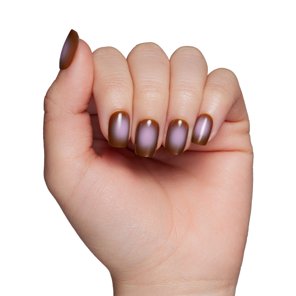 A hand with slightly curled fingers displays short squoval nails painted in a taupe brown and purple metallic gradient from the Eclipse set by glamnetic, shown against a white background.