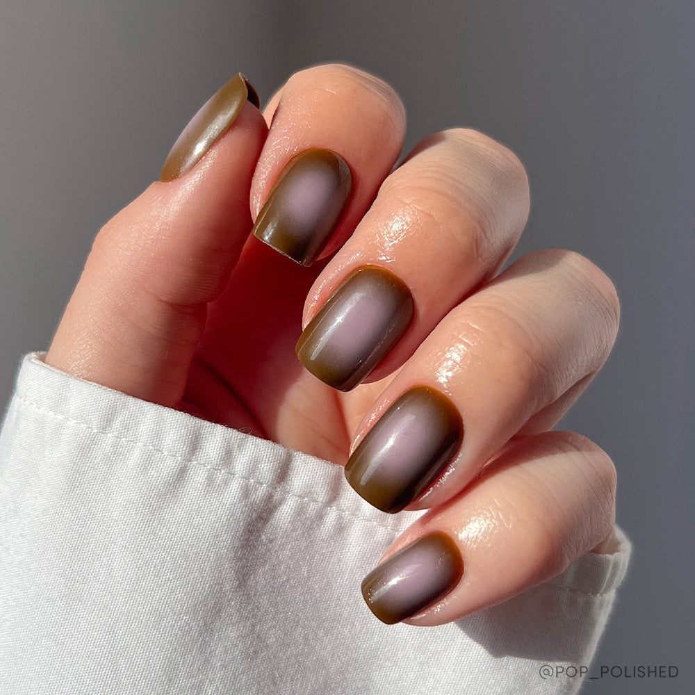 A hand with short squoval nails shows off glamnetics Eclipse, an ombre matte manicure in olive green and taupe brown. A white sleeve and sunlight highlight the chic, well-groomed look.