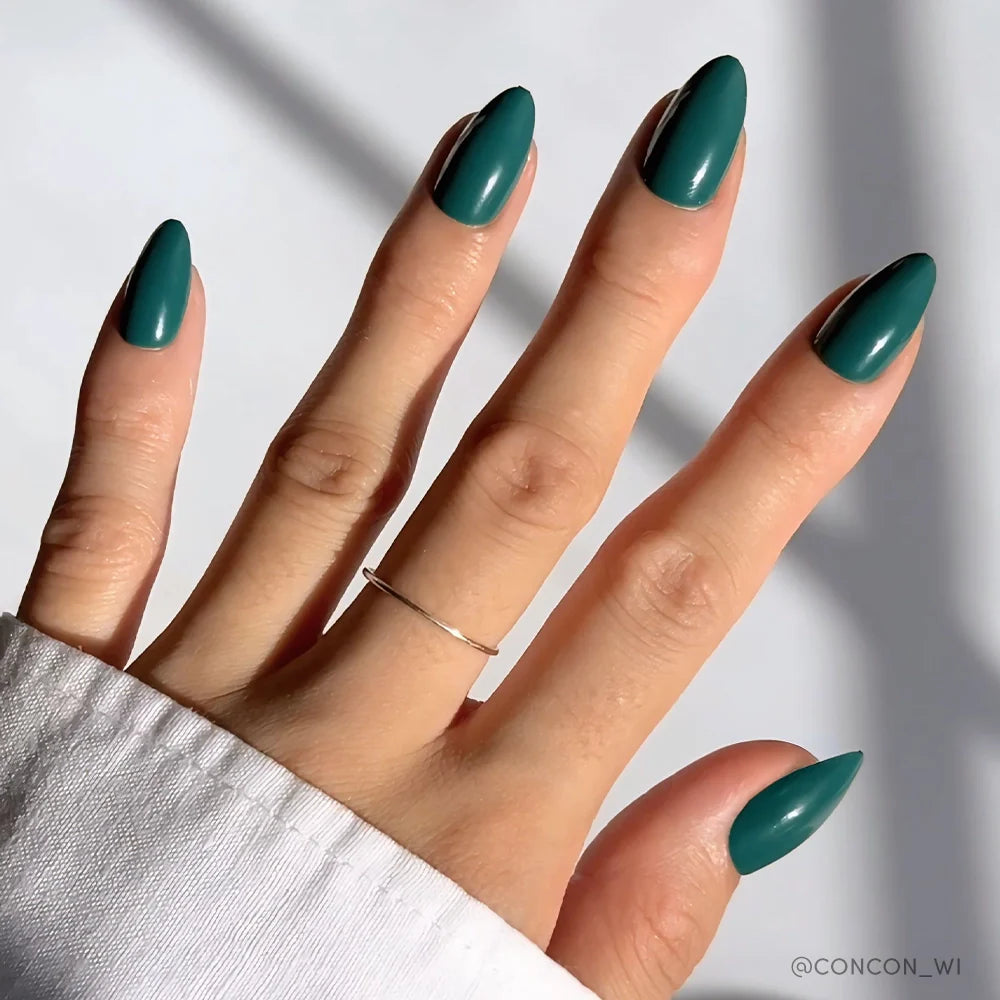 A hand with long, almond-shaped nails features the glossy "Emerald Green" polish by glamnetic against a light background. A simple silver ring on the middle finger and a white cuffed sleeve highlight the vibrant finish.