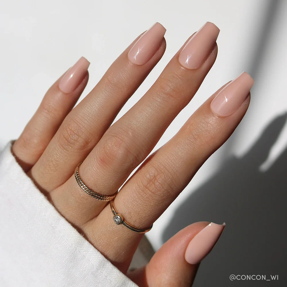 A hand with long, coffin-shaped pinky nude nails in Exposed by glamnetic, featuring a high-shine finish and two gold rings on the middle and ring fingers. A white sleeve and neutral background complete the look.