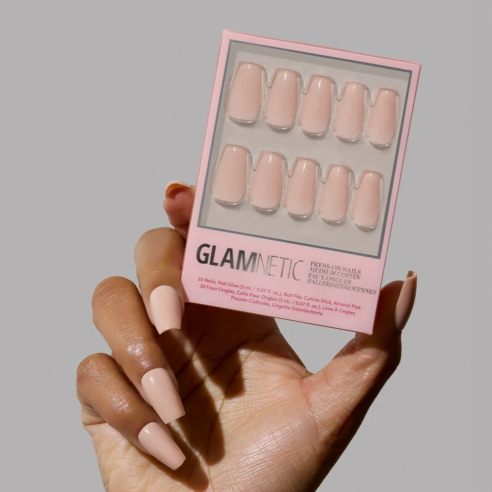 A hand with long, glossy, coffin-shaped glamnetic Exposed press-on nails in a pinky nude shade holds a matching box against a plain gray background.