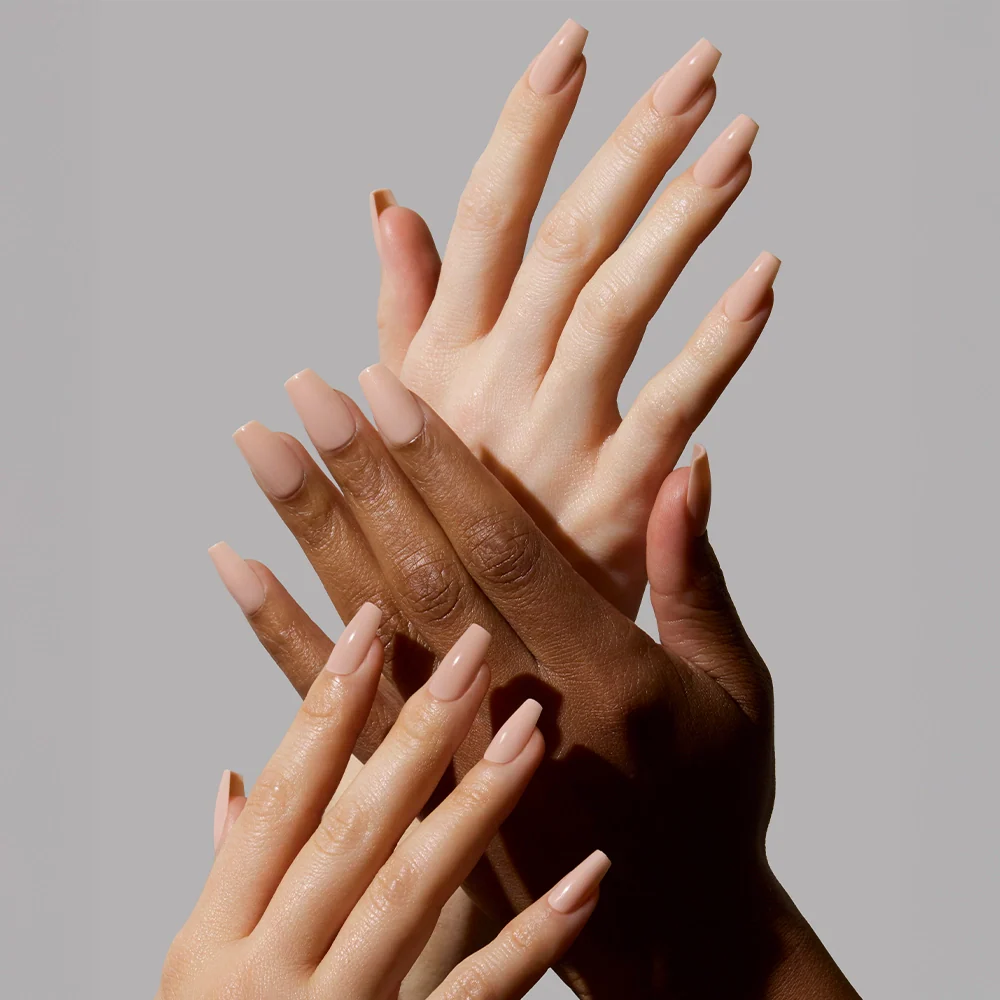 Four hands with different skin tones display long, coffin-shaped nails painted in glamnetic’s Exposed, a pinky nude polish. The overlapping pose highlights the high-shine finish against a plain gray background.