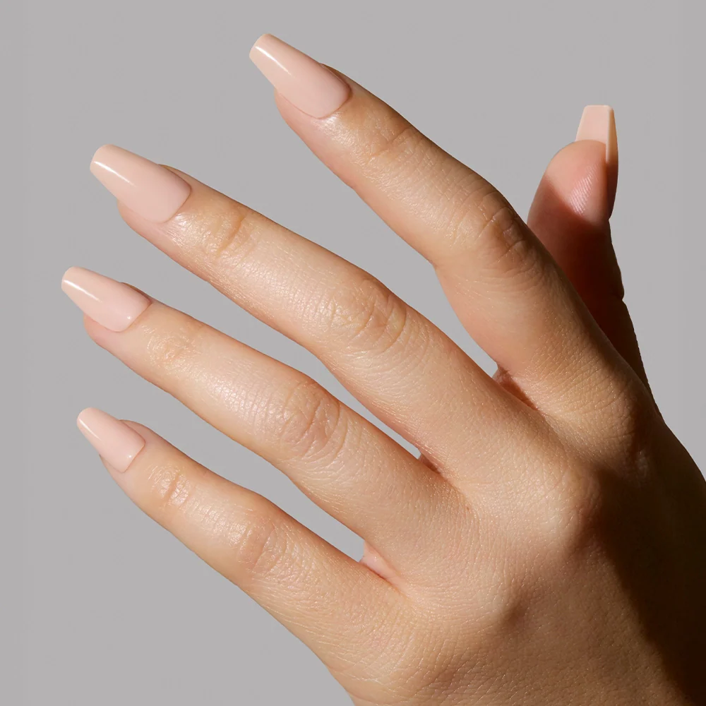A hand with medium-length, square nails painted in glamnetics Exposed, a pinky nude shade with a glossy, high-shine finish, is shown against a plain light gray background.