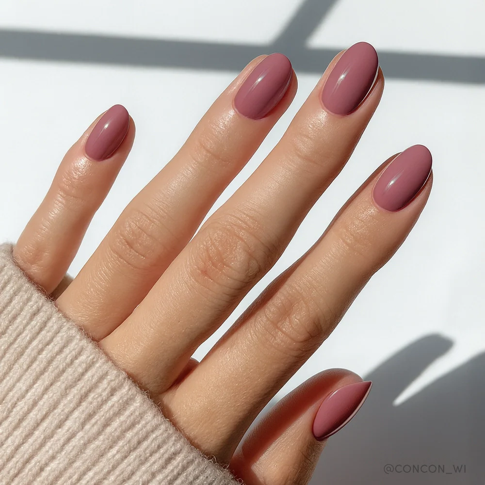 A hand with glamnetic’s Fall Mauve oval press-on nails, featuring a glossy mauve shade, peeks out from a beige ribbed sweater sleeve against a soft, sunlit background—perfect for embracing cozy fall nail trends.