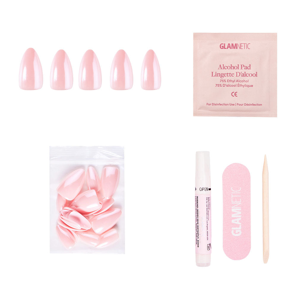 The First Kiss nail set by glamnetic includes blush pink nails, extra nails, nail glue, a pink file, a wooden cuticle stick, and an alcohol pad—all neatly displayed on a white background.