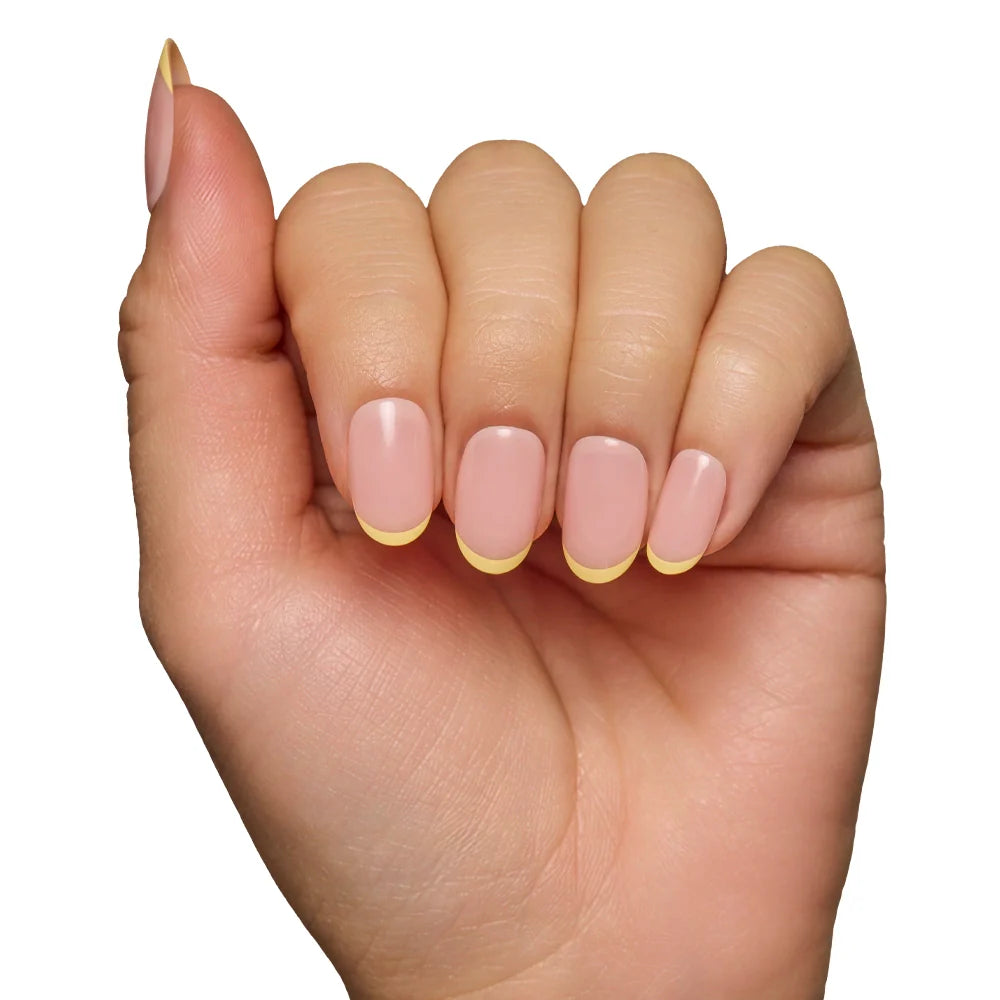 A hand with fingers curled inward displays salon-quality durability and neatly manicured nails from the glamnetic French Butter set, featuring a nude base and pastel yellow French tips against a white background.