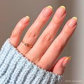 A neatly manicured hand features pastel yellow French tips from the glamnetic French Butter Press-On Nail Set, with a thin gold ring and a light blue ribbed sweater sleeve partially covering the hand.