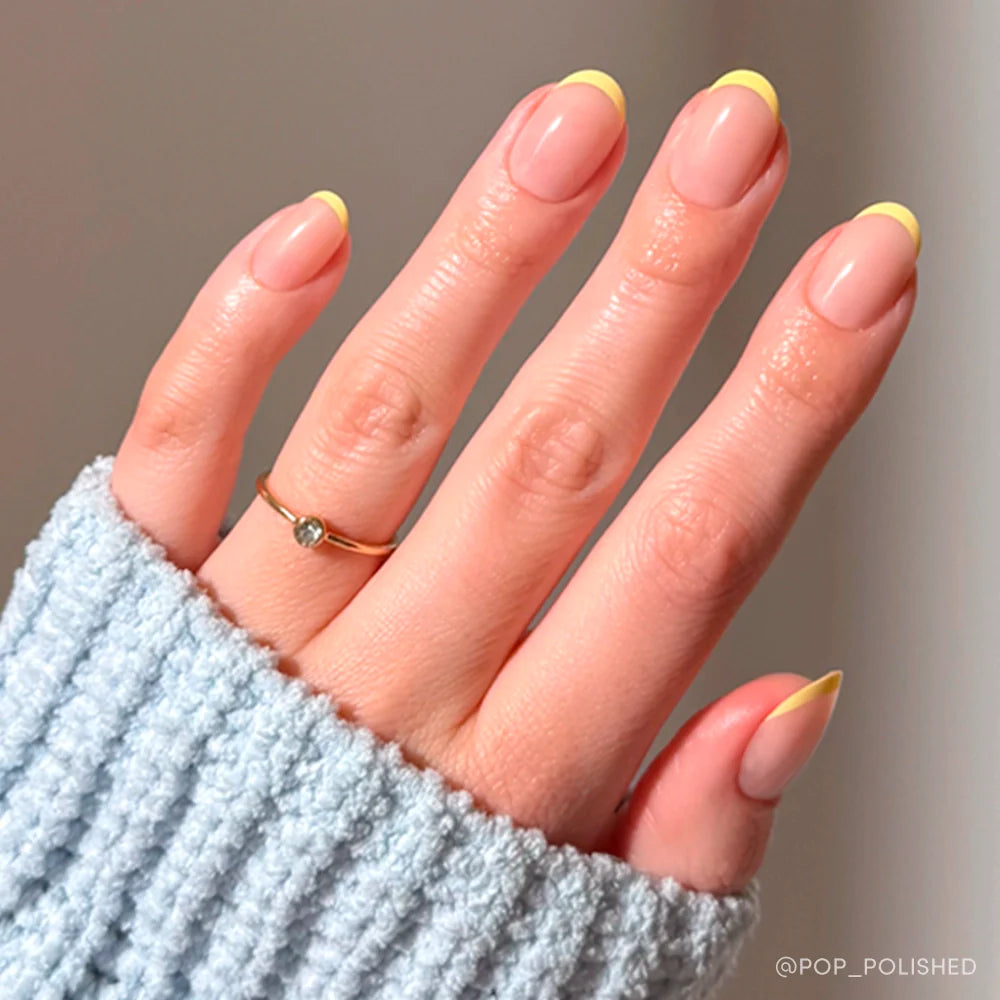 A neatly manicured hand features pastel yellow French tips from the glamnetic French Butter Press-On Nail Set, with a thin gold ring and a light blue ribbed sweater sleeve partially covering the hand.