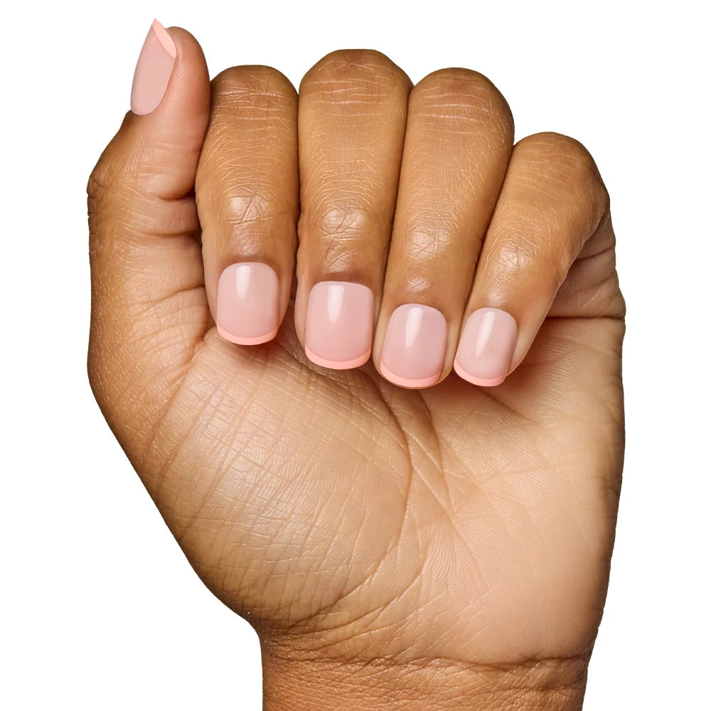 A hand with fingers curled inward displays glamnetics French Peach Press-On Nails, featuring a super short, light pink design. These vegan and cruelty-free nails offer a neat, elegant look against a white background.