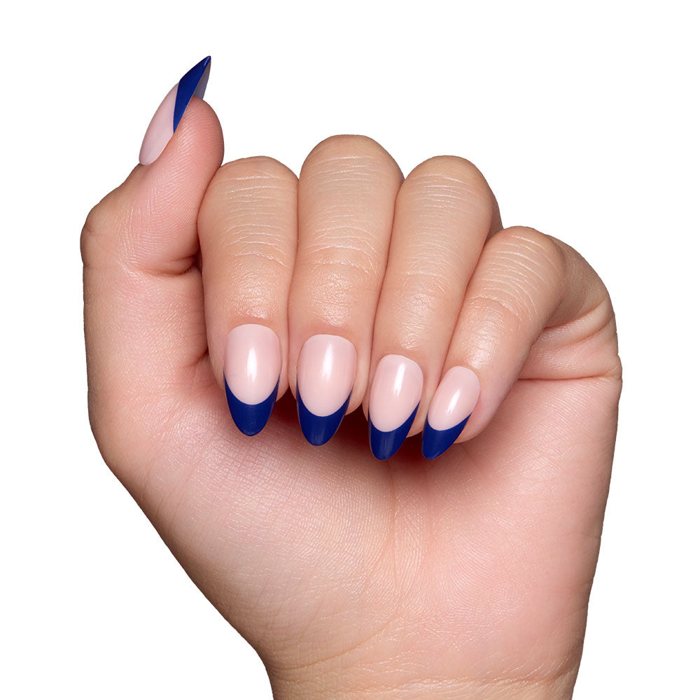 A hand with short almond nails, styled with glamnetics French Riviera—nude base and elegant navy French tips—is shown against a white background.