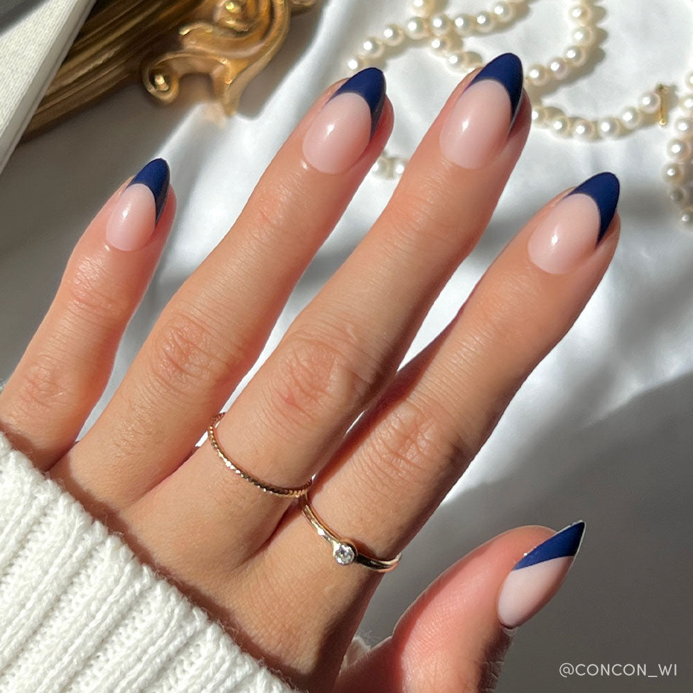 A hand with short almond nails in glamnetics French Riviera—nude base and navy French tips—wears two gold rings and a white knit sweater, with pearls and gold accents in the background.
