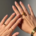 Close-up of hands wearing glamnetic Glam Goals short oval press-on nails in nude pink with gold crescent accents, paired with chunky gold rings and a bracelet for a chic finish.