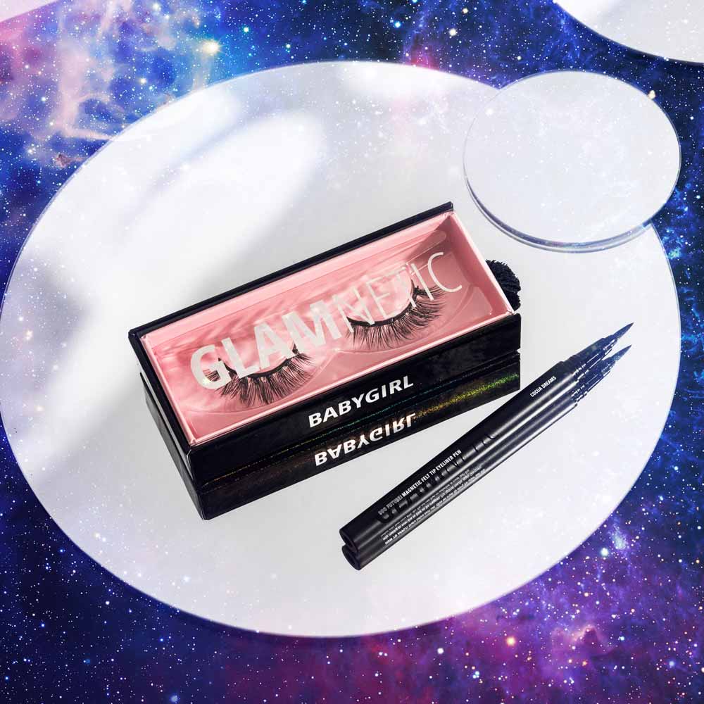 A box of glamnetic Babygirl vegan false eyelashes and two black liquid eyeliners are displayed on a mirrored surface with a galaxy background. These reusable lashes create bold, glamorous looks.