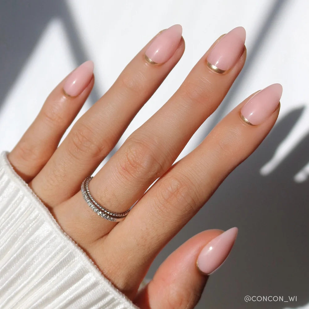 A hand with short almond nails in glamnetic’s Goal Chaser, a light pink polish with minimalist gold half-moon and French tip designs. Two thin, stacked rings with small gems are worn. The white ribbed sleeve completes the softly lit look.
